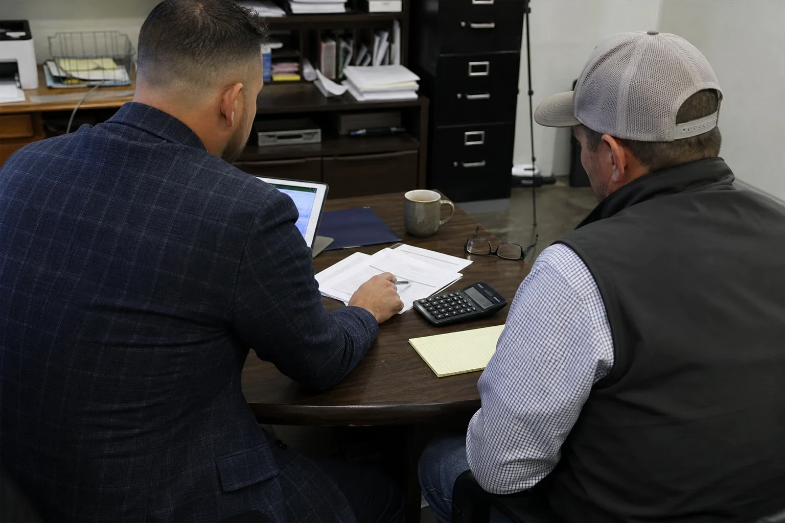 Agent in a plaid blazer reviews policy papers with a business owner in a cap and vest at a wooden office table with a calculator, notepad, laptop and coffee mug