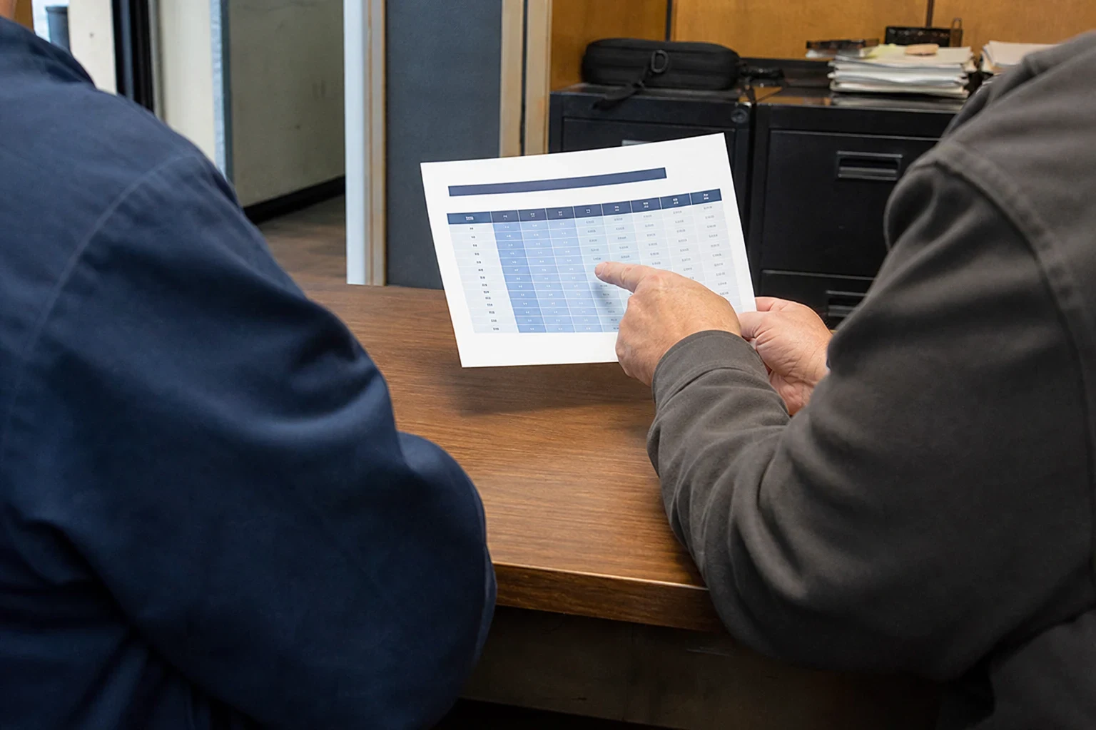 Over-the-shoulder view of a man in a gray shirt pointing to a printed coverage table while another man in navy looks on at a wooden desk in a back office