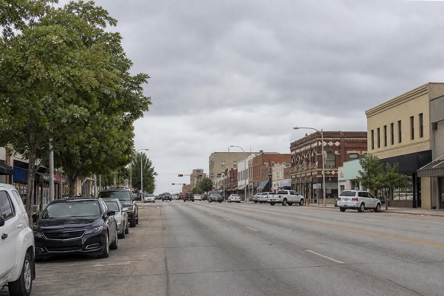Wide downtown main street under overcast skies, with cars parked along both curbs, leafy trees on the left and brick storefront buildings lining the road.